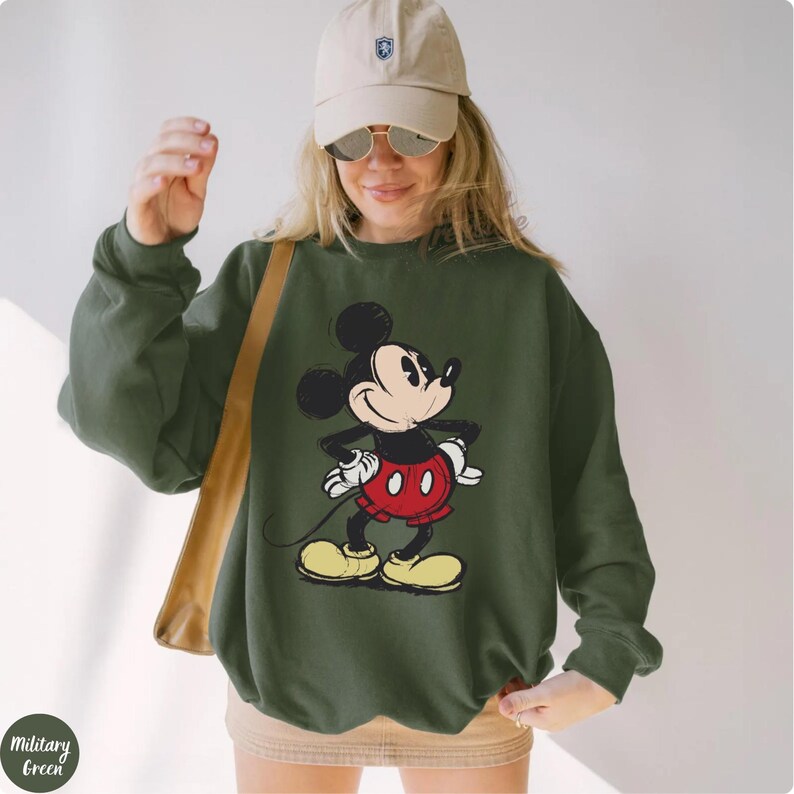 Pode incluir: Camisola verde militar com um gr&aacute;fico desgastado do Mickey Mouse. A personagem de desenho animado usa suas cl&aacute;ssicas cal&ccedil;as vermelhas, sapatos amarelos e luvas brancas. A camisola tem gola redonda e mangas compridas.