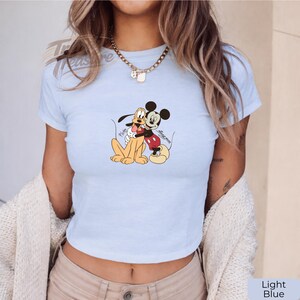 Puede incluir: Top corto azul claro con un gr&aacute;fico de Mickey Mouse abrazando a Pluto. Los personajes de dibujos animados est&aacute;n en el centro de la camiseta. La camiseta es de manga corta y tiene cuello redondo. Las palabras "Pluto" y "Mickey Mouse" est&aacute;n escritas en la camiseta.
