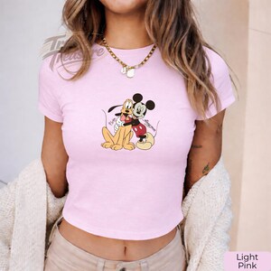 Puede incluir: Top corto rosa claro con un gr&aacute;fico de Mickey Mouse abrazando a Pluto. Los personajes de dibujos animados est&aacute;n a color. La camiseta tiene mangas cortas y cuello redondo. El texto "Mickey & Friends" est&aacute; escrito en la camiseta.