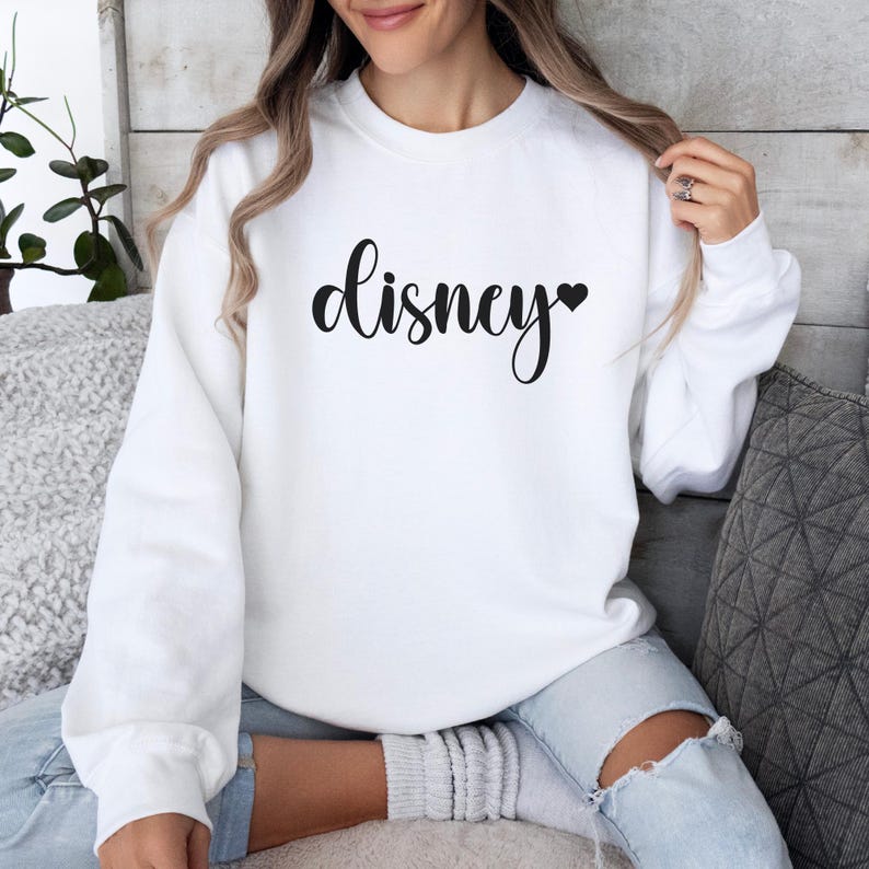 May include: White sweatshirt with the word "Disney" in black script, a black heart replaces the letter "i". The sweatshirt has long sleeves and a crew neck. The model is wearing light blue jeans.