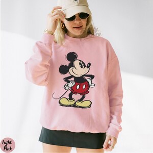 Pode incluir: Camisola rosa claro com um desenho do Mickey Mouse. A personagem de desenho animado &eacute; em preto, vermelho, amarelo e branco. A camisola tem mangas compridas e gola redonda. A imagem &eacute; uma ilustra&ccedil;&atilde;o em estilo vintage.