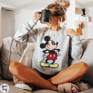 Pode incluir: Camisola cinza claro com um desenho do Mickey Mouse. Mickey &eacute; retratado na sua pose ic&oacute;nica, usando cal&ccedil;&otilde;es vermelhos, sapatos amarelos e luvas brancas. A camisola tem mangas compridas. O texto "Sport Gray" est&aacute; impresso na parte inferior esquerda.