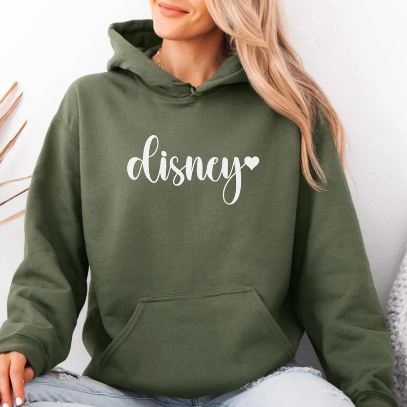 May include: An olive green hooded sweatshirt with a front pocket. The word "Disney" is printed in white script across the chest, with a small heart symbol at the end. The sweatshirt is made of a soft material.