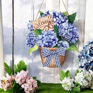 May include: A decorative hanging basket filled with blue hydrangea flowers and green leaves. The basket has a blue and white checkered bow. A small sign with the word "Welcome" is attached. The background is a white wooden fence.