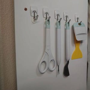 May include: A collection of craft tools, including scissors, a weeding tool, and a scraper, hanging on a white wall. The tools have white handles with mint green accents. A yellow scraper is also included.