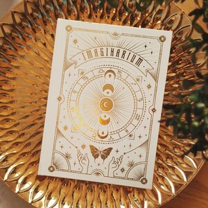 May include: A white journal with gold foil detailing, inscribed with "IMAGINARIUM" and celestial imagery, sits on a gold decorative plate. The plate has a sunburst pattern. The journal cover features moons, stars, and a butterfly design.