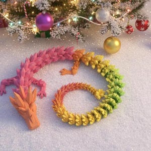 May include: A colorful, articulated dragon toy in shades of pink, orange, yellow, and green, resting on a snowy surface. The dragon is posed near a decorated Christmas tree with ornaments and lights. A festive elf figure and wrapped gifts are also present.
