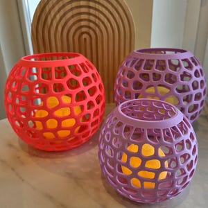 May include: Three decorative sphere-shaped candle holders in shades of red, purple, and lavender. Each holder has a lattice design and contains a yellow light. A wooden arched decor piece is in the background.