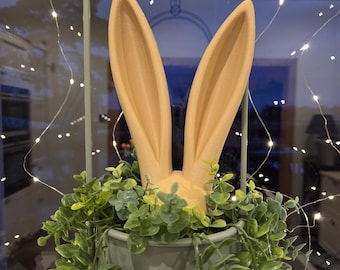 May include: A decorative Easter bunny ears and head, in a light peach color, emerging from a gray pot filled with green foliage. The pot is suspended from a metal frame. String lights are in the background.