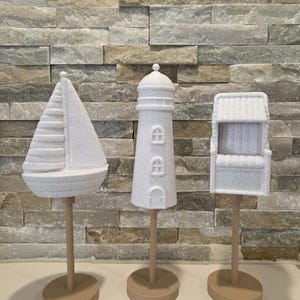 May include: Three white nautical ornaments: a sailboat, a lighthouse, and a small building. Each is mounted on a beige stand with a circular base. The objects have a textured, speckled appearance, suggesting a stone or concrete material.
