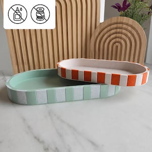 Könnte beinhalten: Zwei ovale Tabletts mit abwechselnd farbigen und strukturierten vertikalen Paneelen. Ein Tablett ist hellgrün und weiß, das andere orange und weiß. Ein weißes Schild darüber zeigt "kein kleber und keine ams" mit Symbolen für keinen Kleber und keinen Faden.