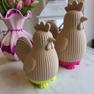 Könnte beinhalten: Zwei beige Hahnenfiguren mit strukturierten Körpern und geschlossenen Augen. Einer hat einen neongelben Sockel, der andere einen rosa Sockel. Im Hintergrund eine rosa Vase mit lila Akzenten und rosa Tulpen.