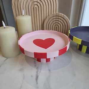 Könnte beinhalten: Dekoratives rosa Tablett mit rotem Herzdesign und rot-weiß kariertem Rand. Das Tablett steht auf einem Sockel. Zwei cremefarbene Kerzen und Holzbogendekorationen befinden sich im Hintergrund.
