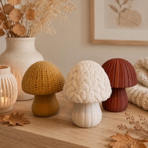 May include: Three mushroom-shaped decorative objects with textured caps. One cap is mustard yellow with a knit-like pattern, one is off-white with a braided cable knit texture, and one is a solid deep terracotta red with vertical ribbed detailing.