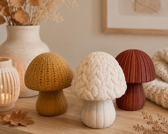 May include: Three mushroom-shaped decorative objects with textured caps. One cap is mustard yellow with a knit-like pattern, one is off-white with a braided cable knit texture, and one is a solid deep terracotta red with vertical ribbed detailing.