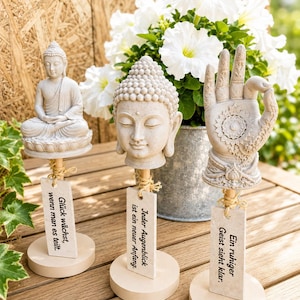 May include: Three decorative stone-coloured ornaments on wooden stands. One features a seated Buddha, one a Buddha head, and one an open hand with a mandala design. Each includes a rectangular tag with German text tied with twine.