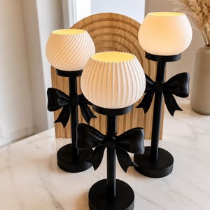 Könnte beinhalten: Drei Tischlampen mit mattschwarzen runden Sockeln, schlanken schwarzen Stielen und dekorativen schwarzen Schleifen an den Stielen. Jede Lampe hat einen weißen, kugelförmigen, gerippten Glasschirm, der ein warmes, sanftes Licht ausstrahlt.