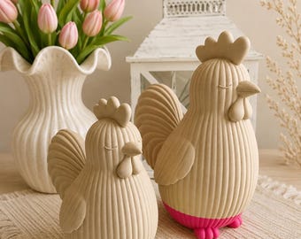 May include: Two beige ceramic chicken figurines with vertical ribbed textures and closed eyes. The smaller chicken has bright neon yellow feet and base, while the larger chicken features vibrant hot pink feet and base, standing on a woven mat.