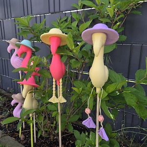 Könnte beinhalten: Eine Gruppe von bunten Flamingo-Gartensteckern, die Hüte tragen. Die Flamingos sind in Rosa, Gelb und Lila gehalten. Die Pfähle sind in einem Gartenbeet mit grünen Blättern platziert.