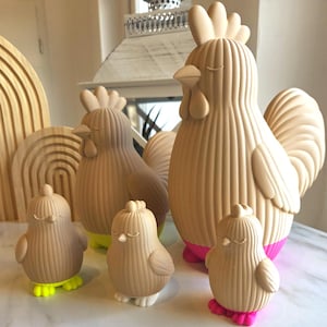 Puede incluir: Una colección de figuras de gallos y pollitos beige con líneas verticales texturizadas. Las figuras de gallo tienen bases rosas, mientras que los pollitos tienen bases amarillas, blancas y rosas. Las figuras tienen los ojos cerrados, sugiriendo un tema pacífico.