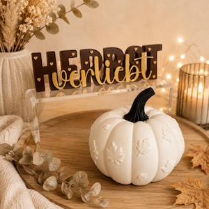 Può includere: Una zucca decorativa bianca con gambo nero e motivi a foglia in rilievo, posizionata accanto a un cartello marrone scuro con testo dorato "HERBSTverliebt" e piccoli cuori dorati, su un vassoio rotondo in legno con fogliame essiccato.
