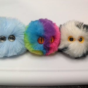 May include: Three plush toys shaped like balls with googly eyes. The first toy is light blue, the second is rainbow colored, and the third is black and white.
