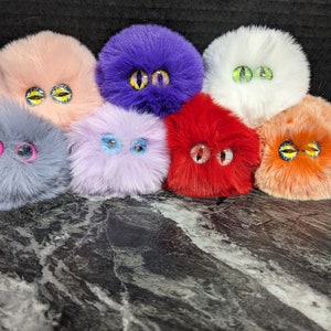 May include: Eight fluffy, round plush toys with googly eyes in various colors, including purple, pink, gray, white, light blue, red, and orange.