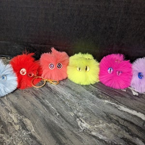 May include: Six colorful fluffy keychains with googly eyes. The keychains are in shades of blue, red, pink, yellow, and purple. Each keychain has a gold ring attached.