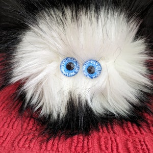 May include: A white and black fluffy toy with blue eyes. The toy is sitting on a red knitted surface.
