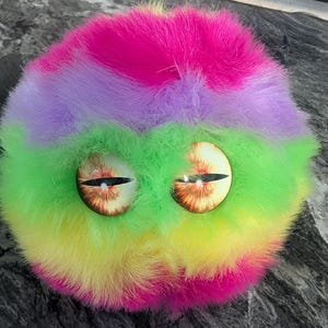 May include: A colorful, fuzzy creature with large, detailed eyes. The plush toy features bands of pink, purple, green, and yellow. The eyes have a brown iris with a black slit pupil, giving it a whimsical appearance.