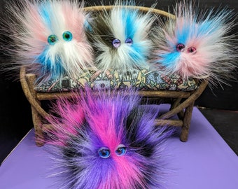 May include: Four fluffy, colorful, fuzzy creatures with googly eyes sitting on a wooden bench. The creatures are made of different colors of fur, including pink, blue, white, and black.