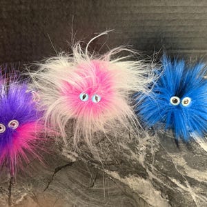 May include: Three fluffy, round, handmade creatures with faux fur in purple, pink, and blue. Each has two googly eyes. The creatures are on a gray and black textured surface.