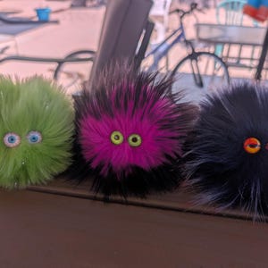 May include: Three fluffy, round, monster-like toys with googly eyes. One is lime green, one is black and pink, and the last is black with orange and yellow eyes. The toys are sitting on a brown surface.