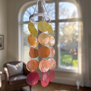 May include: A hanging mobile featuring iridescent, circular shell pieces in shades of yellow, orange, and red. The shells are connected by small metal rings and suspended from a white rope ring. The mobile hangs in front of a window.