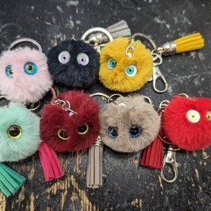 May include: A set of eight colorful fluffy keychains with googly eyes. Each keychain features a different color and has a small tassel attached.