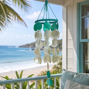 May include: A hanging wind chime with a turquoise and white color scheme. The chime features a turquoise ring at the top, with turquoise and white shell-shaped pieces hanging below. The background shows a beach scene with blue water and sky.