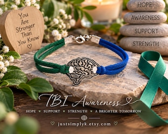 May include: A bracelet featuring a silver-toned brain charm centered between two strands of green suede cord and two strands of blue suede cord, secured with a silver-toned lobster clasp. The bracelet is displayed on a textured stone surface.