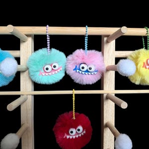 May include: Five fuzzy, round pom-pom keychains in blue, mint green, pink, yellow, and red. Each features two large white googly eyes, a small white pom-pom, and a plastic mouth with white teeth, suspended by a matching colored ball chain.