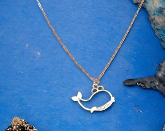 May include: A silver whale pendant necklace on a silver chain. The necklace is displayed on a blue background with rocks.