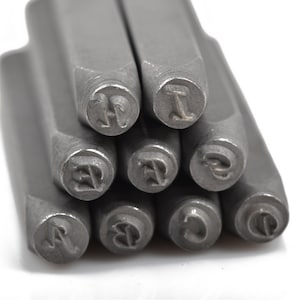 May include: Close-up of a set of metal letter stamps. The stamps are arranged in a cluster, with the lettered ends facing the viewer. The letters visible are A, B, C, D, E, H, and I. The stamps are made of a silver-colored metal.