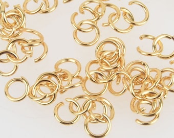 May include: A collection of gold-colored jump rings, scattered across a white surface. The jump rings are open, with a small gap, and appear to be made of metal. These jewelry findings are used for connecting jewelry components.