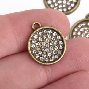 May include: Three round, bronze-colored pendants with a loop at the top. Each pendant is covered with small, clear crystals. One pendant is held in a person's hand, and two are in the background.