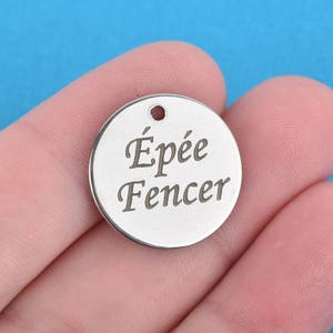 May include: A small, round, silver-colored charm with the words "Épée Fencer" engraved on its surface. The charm has a small hole at the top for attaching to a necklace or bracelet. The background is a solid blue color.