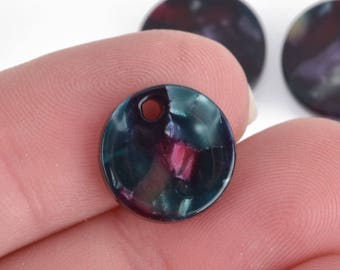 Puede incluir: Primer plano de una cuenta redonda y plana con un agujero en la parte superior. La cuenta es una mezcla de colores verde azulado oscuro, morado y rosa. Otras dos cuentas están ligeramente desenfocadas en el fondo. La cuenta está sostenida por los dedos de una persona.