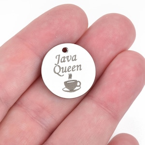 May include: A small, round, silver-colored charm with the words "Java Queen" and a coffee cup design. The charm has a small hole at the top. The charm is held in a person's hand.