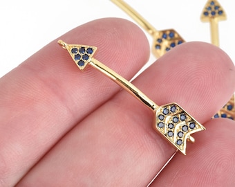 May include: Gold-colored arrow charms with blue gemstone accents. The arrowheads and fletching are adorned with small, dark blue gemstones. The charms are held in a hand against a white background.