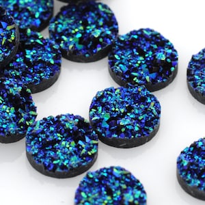 May include: A collection of round, dark blue druzy cabochons with sparkling blue and green crystalline surfaces. These gemstone beads are scattered on a white background, showcasing their unique texture and color variations. Ideal for jewelry making.