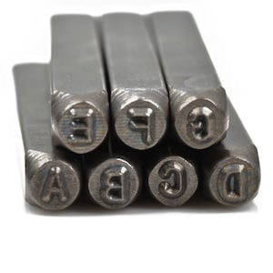 May include: A set of metal letter stamps, featuring the letters A, B, C, D, E, F, and G. The stamps are arranged in a stack, with the lettered ends visible. The metal has a matte finish, and the background is white.