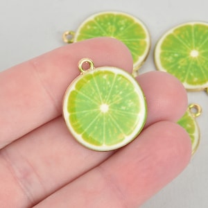 Puede incluir: Primer plano de varios colgantes redondos con forma de rodaja de lima, con un borde dorado y un lazo para sujetarlos a la joyería. Los colgantes tienen un centro verde brillante con detalles blancos, que se asemejan a una lima real. El fondo es gris claro.