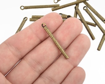 May include: A close-up of a hand holding a gold-toned metal pendant with a textured surface. The pendant is a long, thin rectangle with a loop at the top. There are several other similar pendants scattered around the hand.